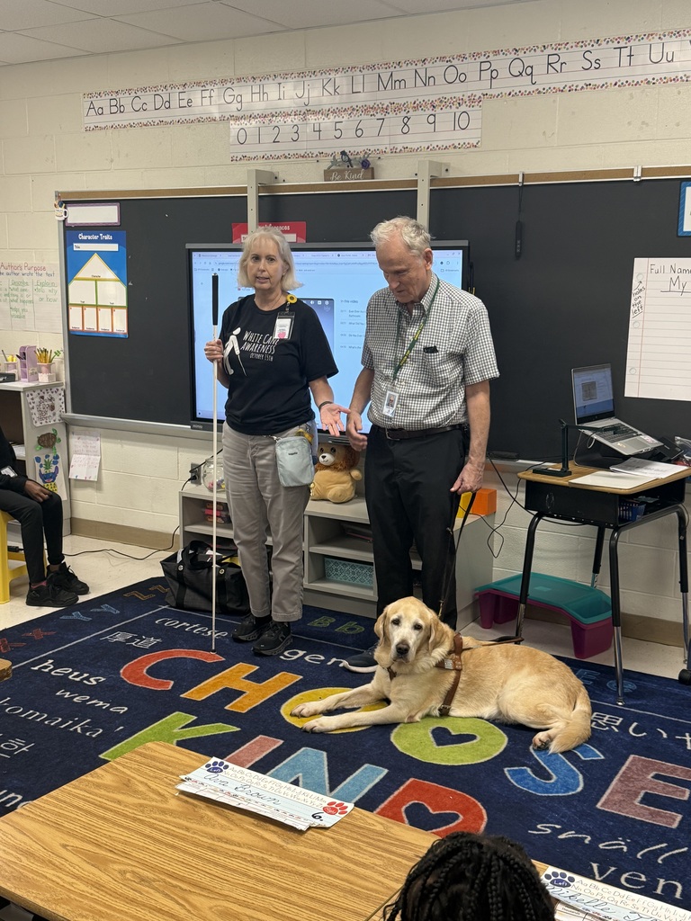 A woman wearing a "White Cane Awareness" t-shirt holds a long white cane while presenting to a classroom alongside a man holding a leash. At their feet, a yellow Labrador retriever guide dog wearing a brown harness rests calmly on a colorful classroom rug with large letters that spell "CHOOSE KIND".