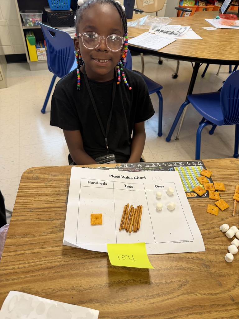 A young female student wearing glasses and hair beads smiles sitting at her desk, where a Place Value Chart worksheet is set up. The chart uses snacks to represent numbers: 1 square cheese cracker in Hundreds, 8 pretzel sticks in Tens, and 4 mini marshmallows in Ones, matching a sticky note below labeled 184.