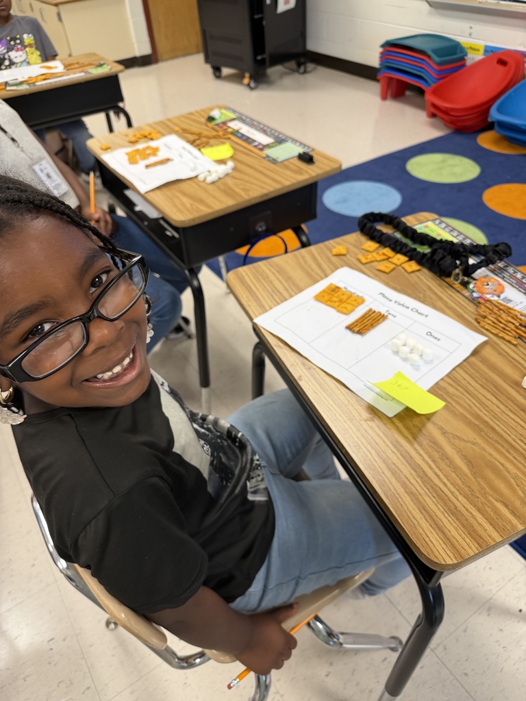A young female student wearing glasses smiles at the camera from her classroom desk. In front of her is a Place Value Chart activity using square crackers, pretzel sticks, and mini marshmallows to represent hundreds, tens, and ones places.