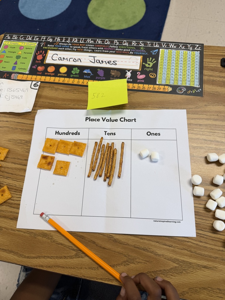 A high-angle view of a student's desk showing a completed Place Value Chart math activity. The sheet displays 5 square crackers under Hundreds, 8 pretzel sticks under Tens, and 2 mini marshmallows under Ones, with a yellow sticky note above showing the target number 582.