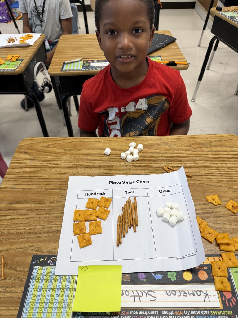 A young male student in a red shirt smiles behind his classroom desk featuring a Place Value Chart worksheet. The chart shows 8 square crackers under Hundreds, 8 pretzel sticks under Tens, and 8 mini marshmallows under Ones, with extra snack supplies and a sticky note labeled 888 on the desk.
