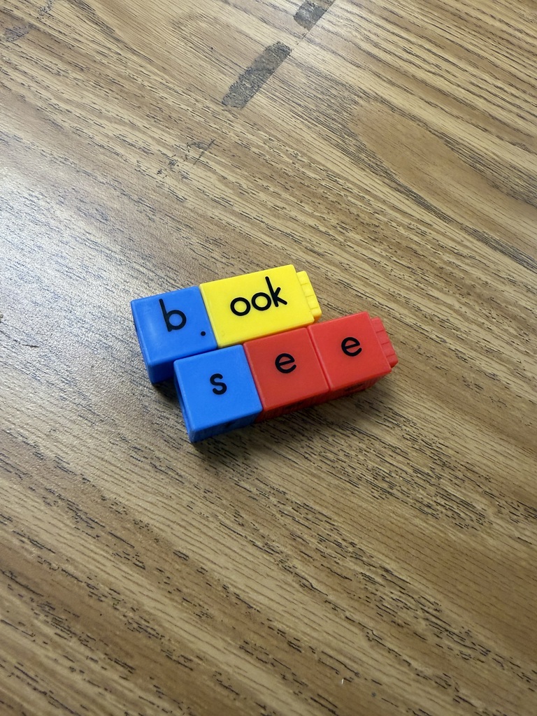 A close-up shot on a wooden tabletop showing interlocking letter blocks snap-connected to spell out words. One set of blue and yellow blocks spells "book," and a connected set of blue and red blocks underneath spells "see".