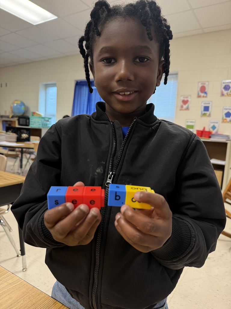 A young student wearing a black jacket holds up connected letter blocks in both hands during a phonics activity. The colorful blocks snap together to create words like "see".