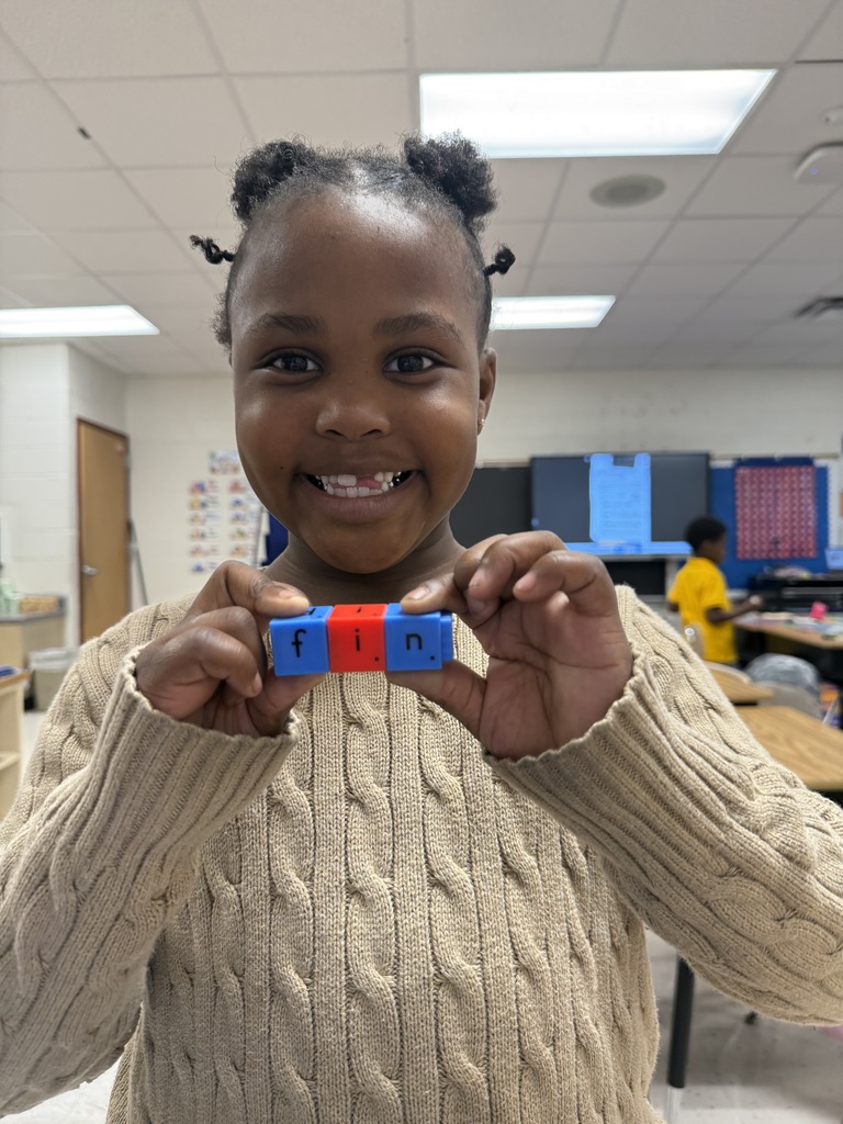 A smiling young female student in a beige cable-knit sweater holds up three snap-together letter blocks in front of her. The blue and red blocks are connected to spell out the word "fin".