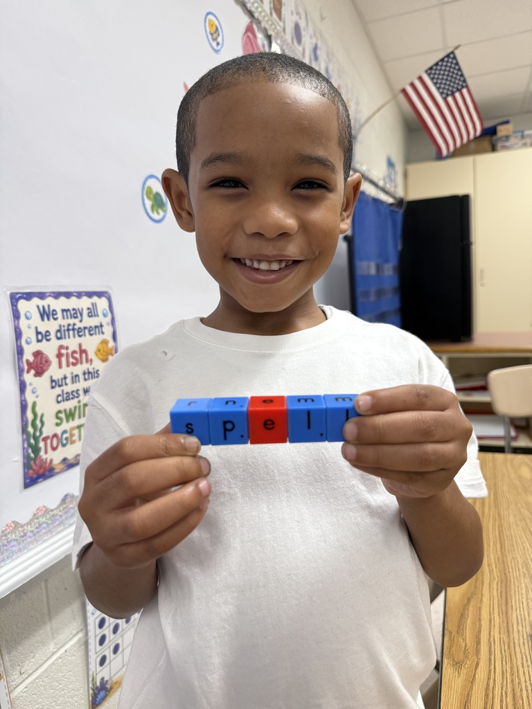 A smiling young male student in a white t-shirt proudly holds up a row of blue and red snap blocks in front of him. The interlocking blocks are arranged to spell out the word "spell".