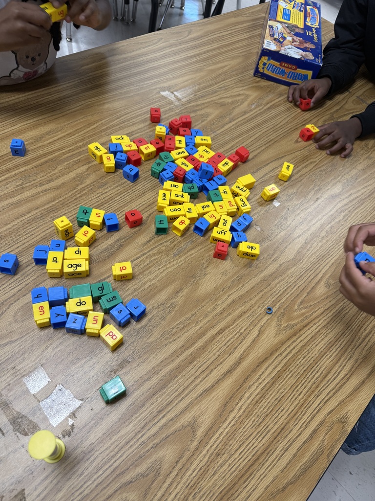 A top-down view of a wooden classroom table scattered with colorful interlocking letter and word-building blocks in blue, red, yellow, and green. Students' hands are visible around the edges of the table as they build words, with a Word-Word game box resting nearby.