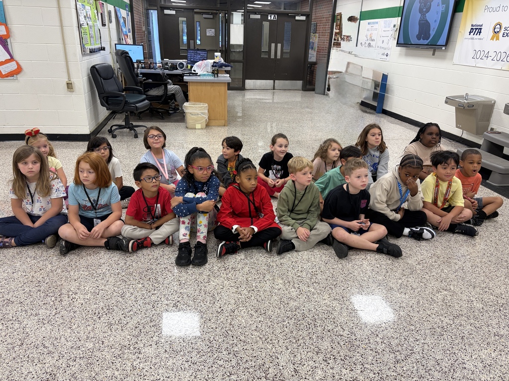 A group of elementary school children sit cross-legged on the floor in a school hallway or lobby, looking attentive. In the background, double doors, a desk setup, and a wall display with a TV screen and National PTA signage are visible.