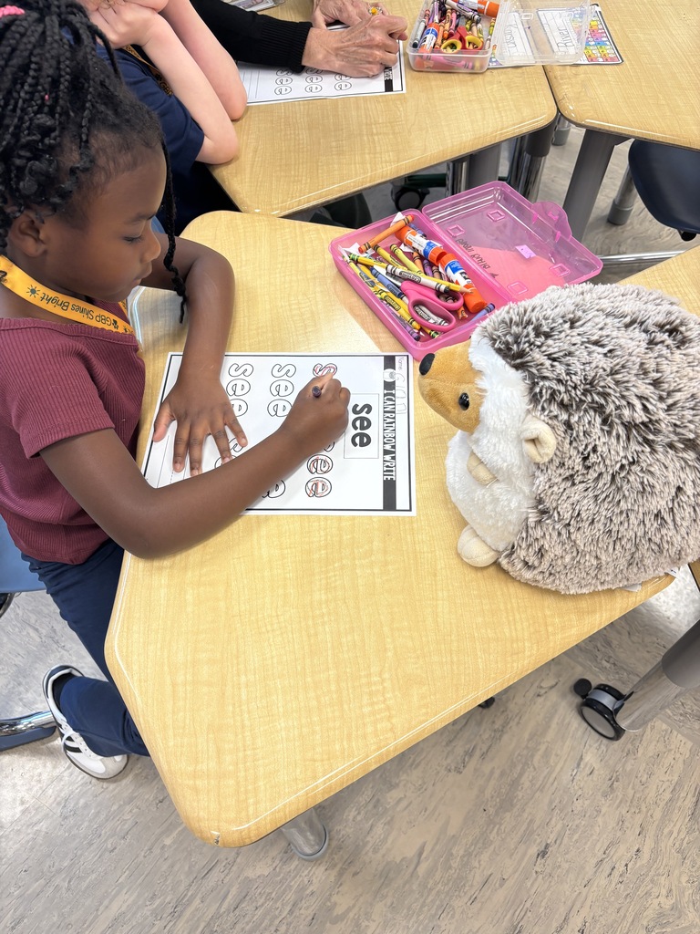 A young girl in a maroon shirt sits at a desk, writing on a paper with crayons.