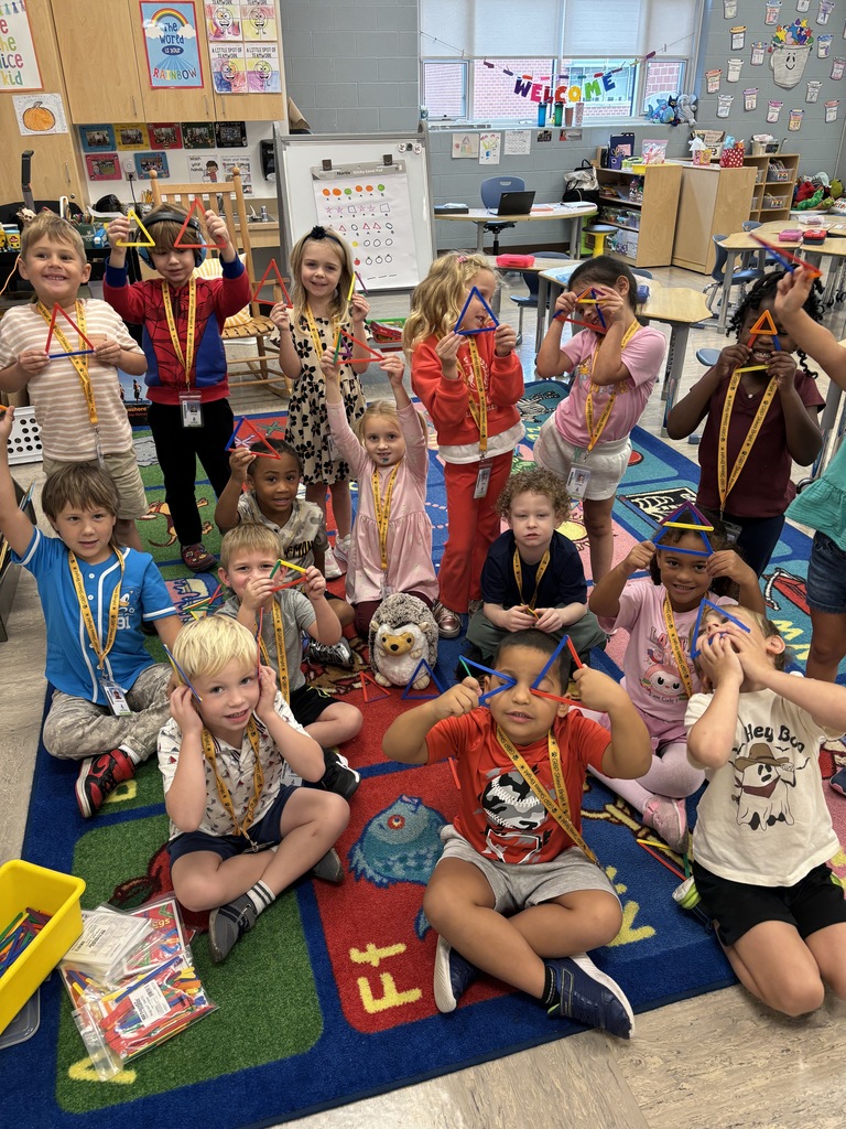 Children sit and stand in a classroom, raising their hands while wearing lanyards, some holding objects.