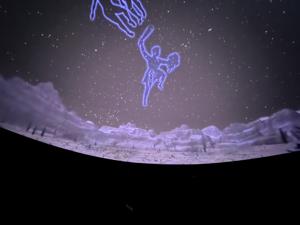 A planetarium dome projection displaying a starry night sky above a rocky landscape, with glowing blue constellation outlines illuminated in the center, including the figure of Orion holding a club and shield.