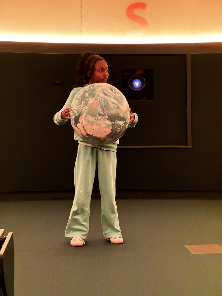 A young girl standing in the front area of a planetarium room, holding a large, lightweight globe model of planet Earth in front of her. She is wearing a light green tracksuit, and a large orange "S" marker is illuminated on the wall above her.