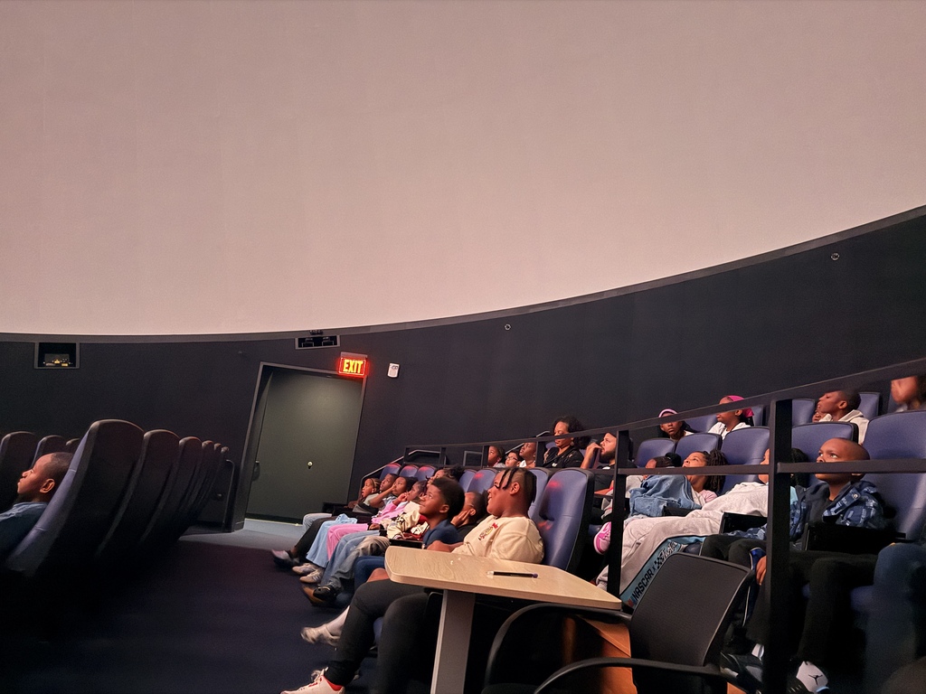 A group of students sitting in comfortable, dark blue theater seats inside a planetarium, all looking upward toward the domed ceiling screen. A glowing red "EXIT" sign is visible near a doorway along the curved wall in the background