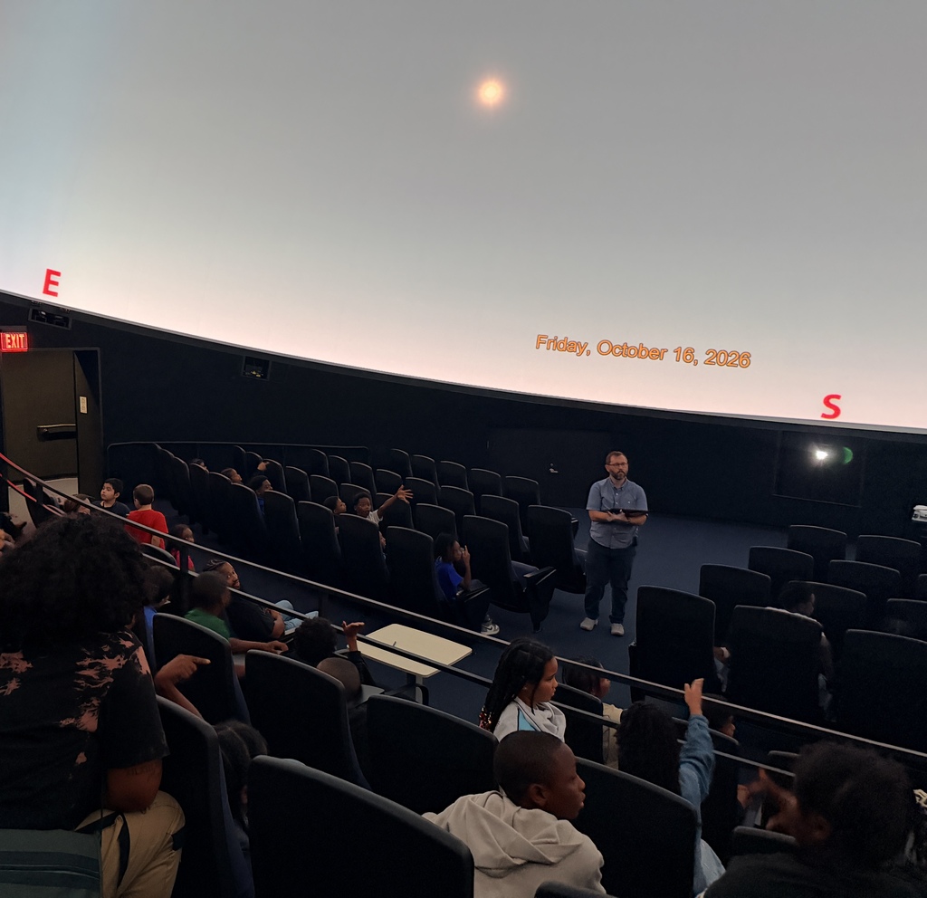 A wide-angle view inside a planetarium showing a group of students seated in tiered rows listening to an instructor standing at the front. The planetarium dome projects a pale sky with a bright light spot and orange text reading "Friday, October 16, 2026", along with red directional markers for "E" (East) and "S" (South). Several students in the foreground have their hands raised.