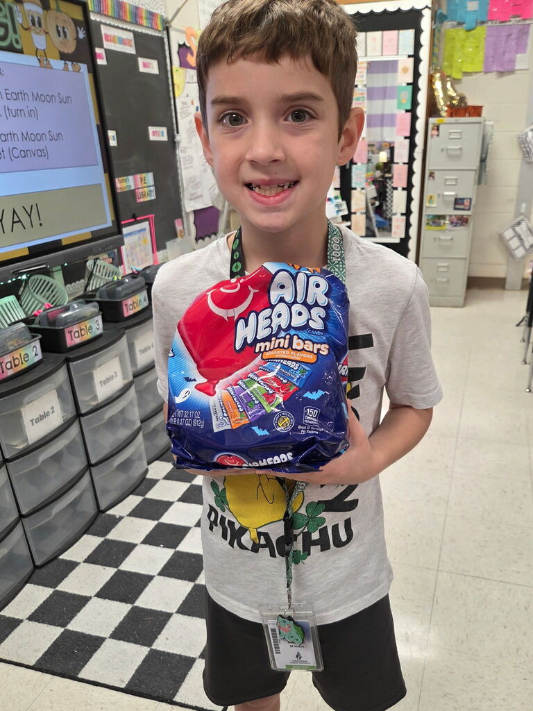 A smiling boy in a classroom holds a bag of Airheads mini bars, with storage units and a black and white checkered floor in the background.