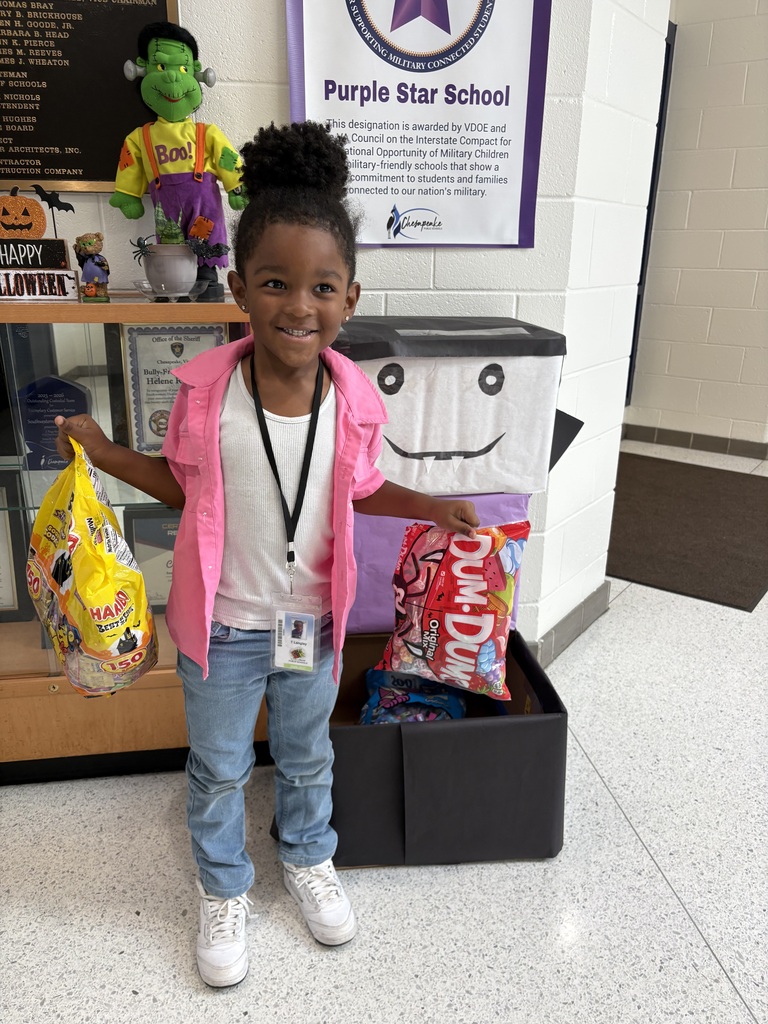 A young girl wearing a pink jacket and jeans stands in front of a box holding a bag of snacks. Behind her is a display case with a green monster, a sign, and other items.