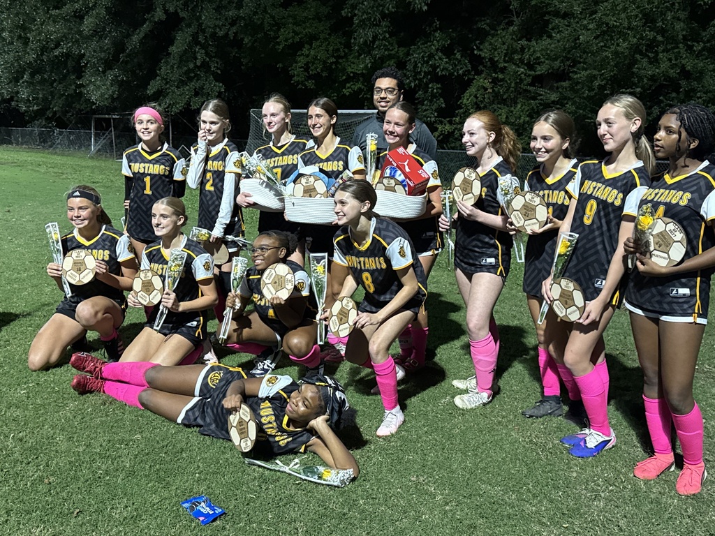 A group of girls wearing pink, black and gold uniforms standing in a group with a coach holding soccer balls.