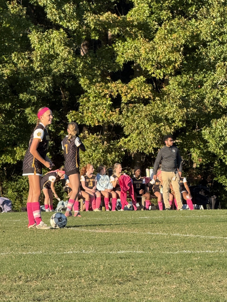 Two girls standing on a soccer field with pink, black, gold uniforms in front of a row of girls seated next to a male wearing brown pants and a black jacket.