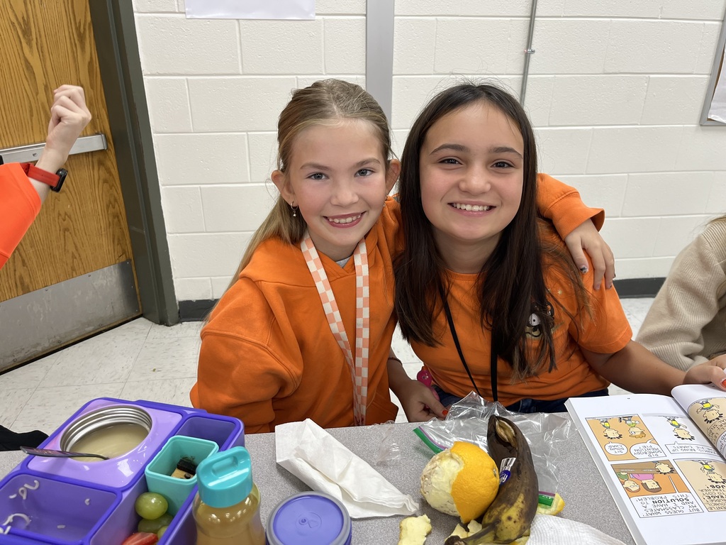 Two students sitting next to each other in the cafeteria.  They are wearing orange shirts.  There is a book and their lunch is on the table in front of them.