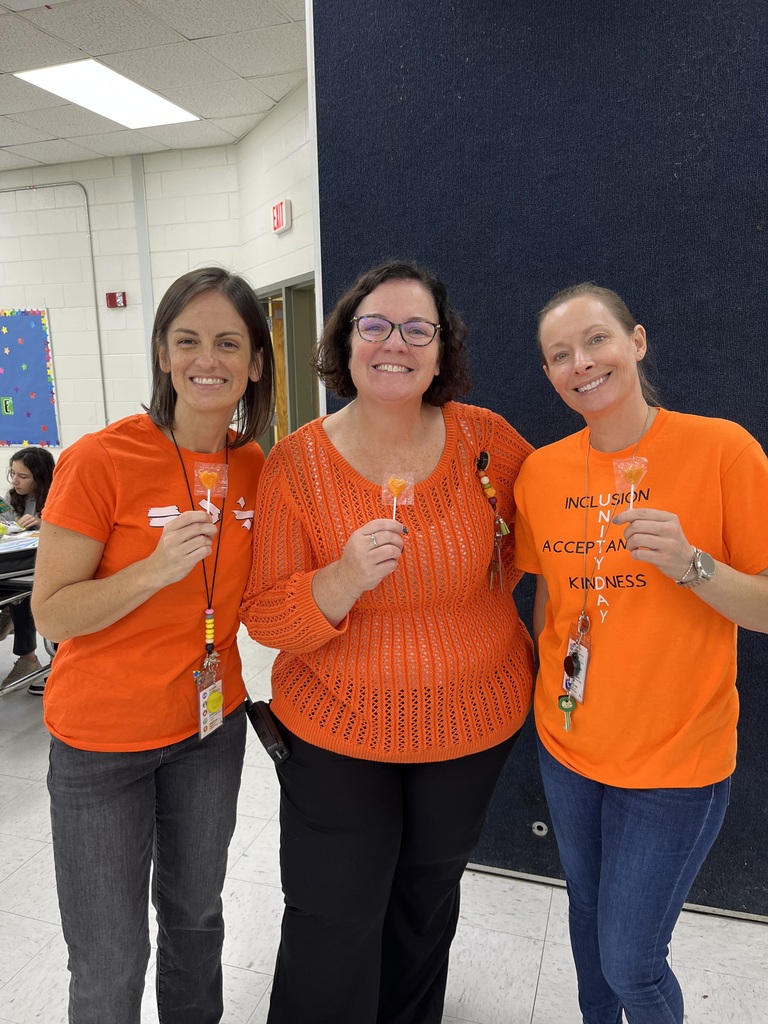 A picture of three adults standing in a room.  They are wearing orange shirts and holding orange lollipops.