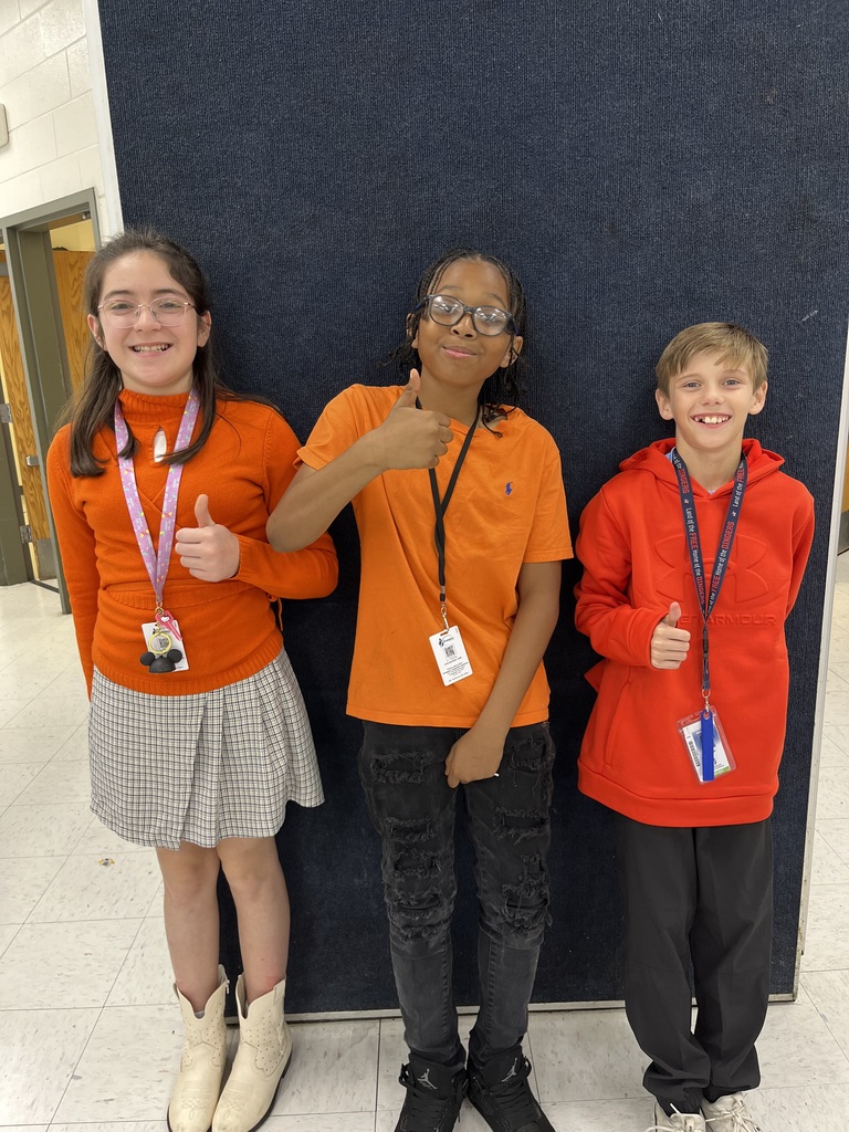 Three kids standing in front of a black background wearing orange shirts.  Two have on black pants and one is wearing a black and white skirt.