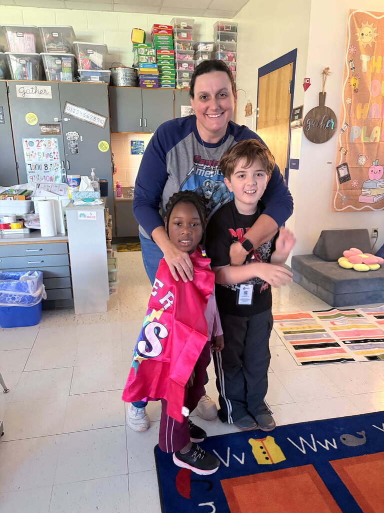 A smiling female teacher wearing a Captain America shirt stands behind two elementary students inside a colorful classroom. One student wears a bright pink superhero cape with a lightning bolt, and the other student wears a black Marvel shirt.