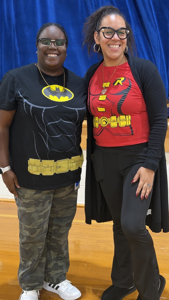 Two female staff members stand side-by-side in a gymnasium in front of a blue curtain, smiling while wearing superhero shirts featuring Batman and Robin logos.