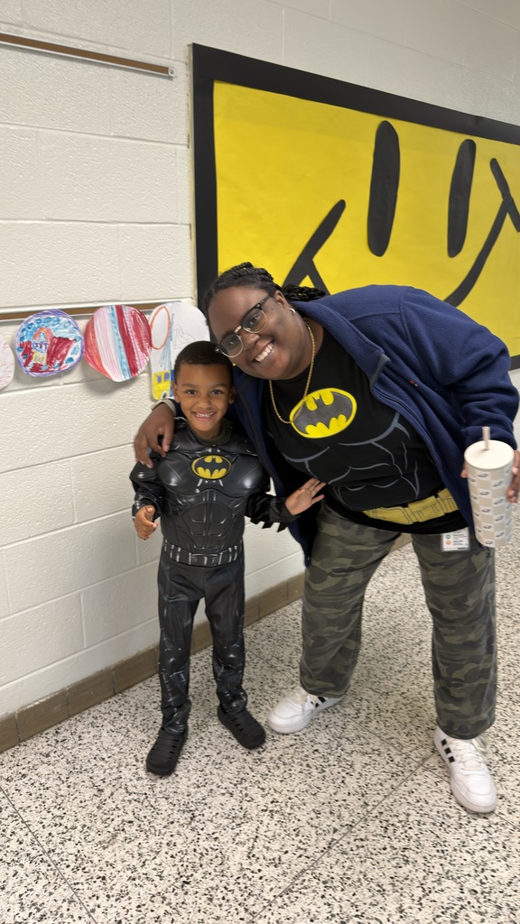 A smiling female staff member in a Batman t-shirt stands next to a young boy student fully dressed in a Batman costume in the school hallway.