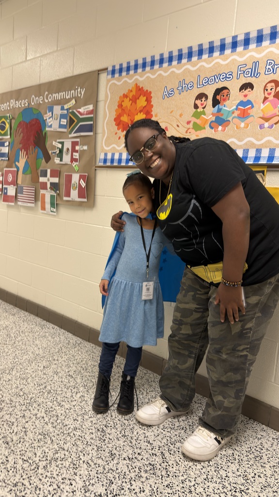 A smiling female staff member wearing a Batman t-shirt and camo pants stands next to a young girl student wearing a blue dress and a matching blue superhero cape in a school hallway.