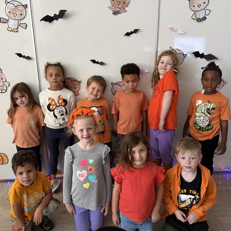 Group of children in orange and other colors standing against a white wall with Halloween decorations