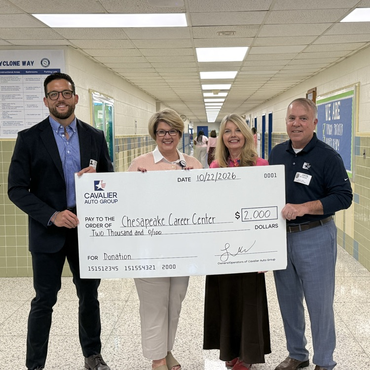 Four people stand behind a large check, holding it up with their hands. Two people wear suits, one wears a dress, and one wears a belt.