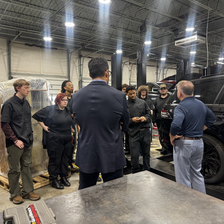 People in a warehouse near a large box, a parked car, and a raised vehicle.