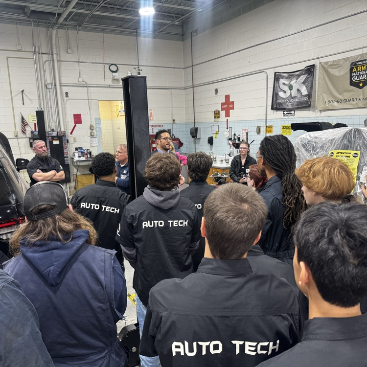Students in black jackets gather around a vehicle in a garage, facing an adult holding a yellow paper.