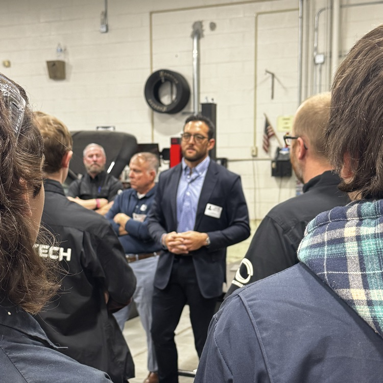 Several adults in jackets and suits stand in a room, some looking at a man in the middle.