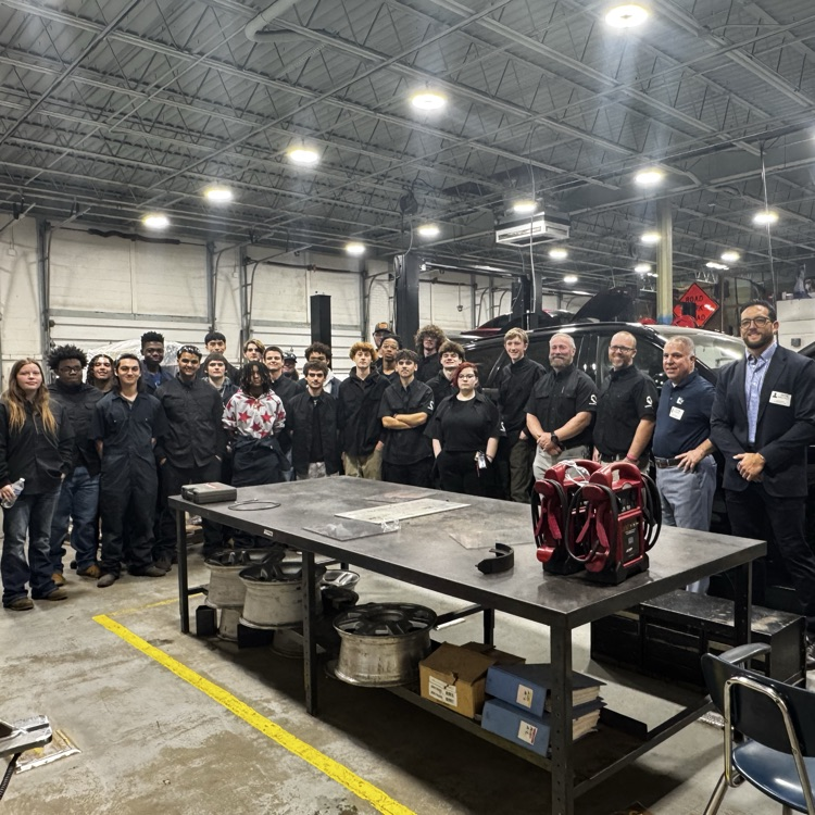 Group of people in a garage; metal tables, a red bag, and other items on a table.