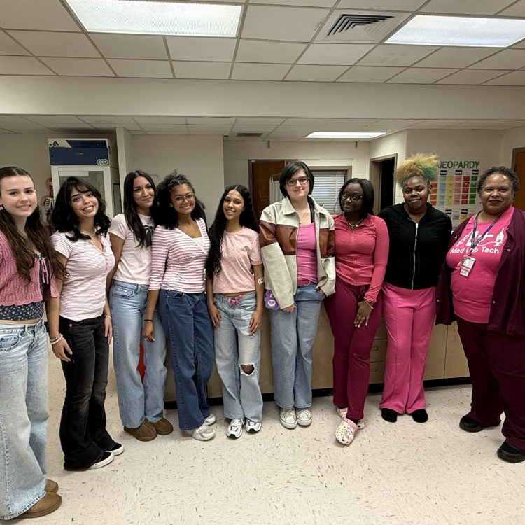 A group of people in pink tops stand in a hallway. Some wear jeans; some wear pants.