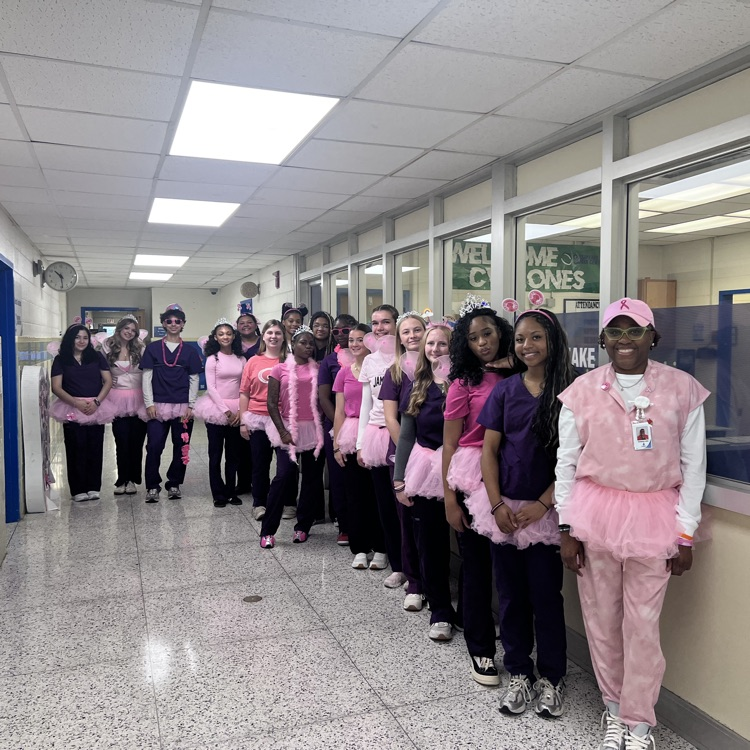 Students in pink tutus stand in a row in a hallway, near windows with green signs.