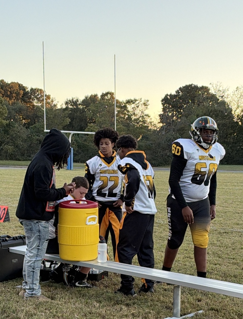A group of football players standing near a water cooler.  They are wearing black, gold and white uniforms.  Another boy wearing jeans and a black shirt is standing with them.