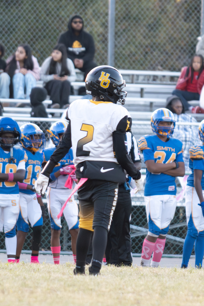 A football player in a black, gold, and white uniform with the number 2 on the jersey is standing facing a line of football players in blue , white and yellow uniforms.