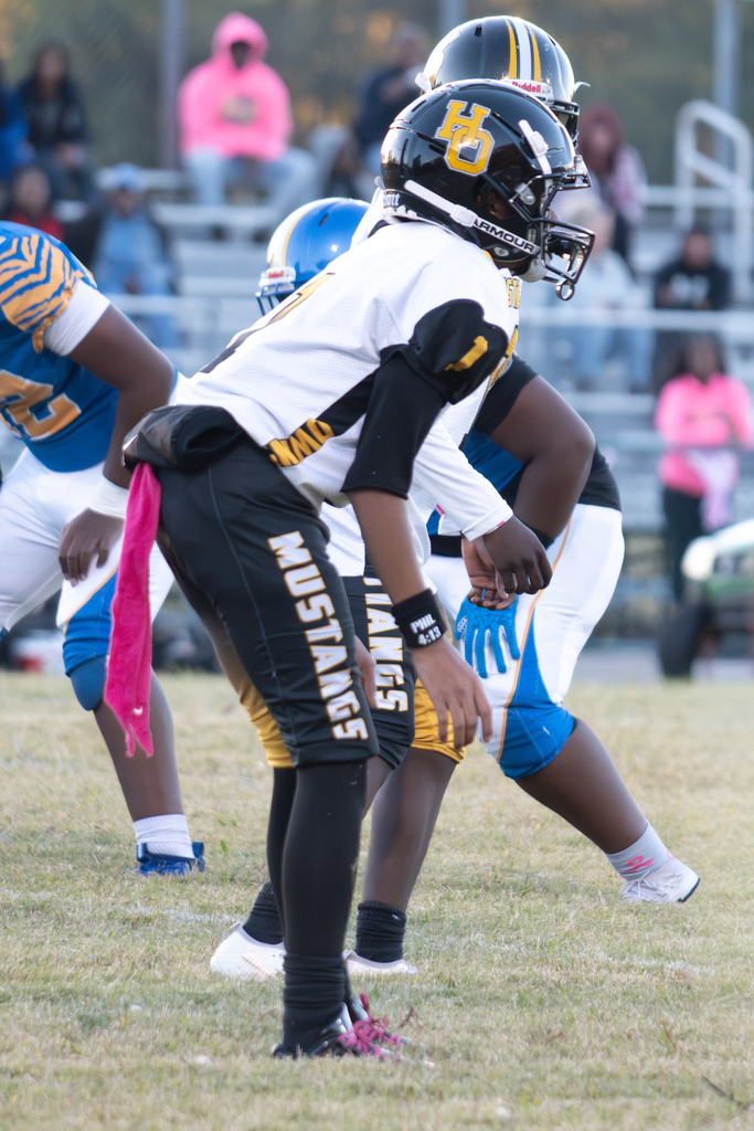 A group of football players are on a field ready to start a play.  Two are wearing black, gold and white uniforms with Mustangs on the pants.   The other 2 are wearing blue and white unforms.