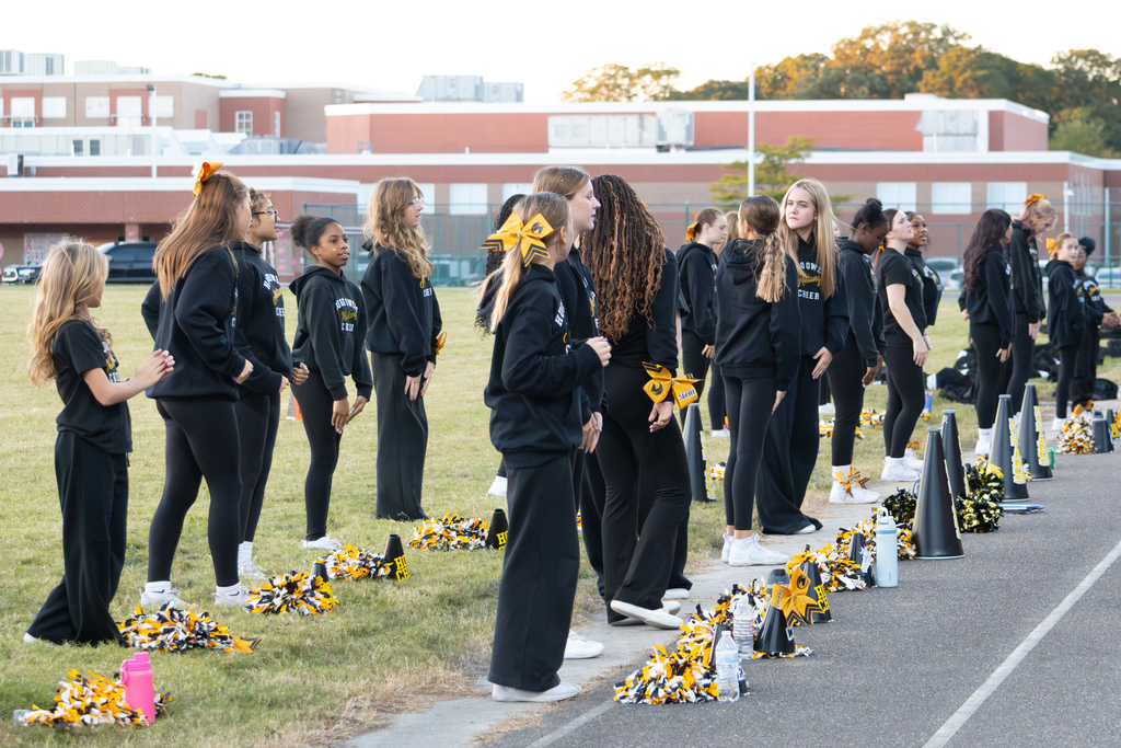 Two rows of cheerleaders are standing with a row of pom poms laying in front of each row.