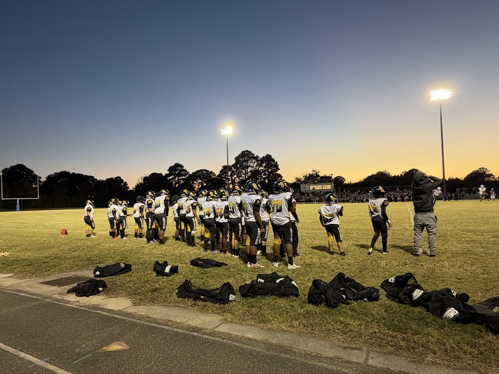 A team of football players standing facing a football field at sunset.  They are wearing black, white and gold uniforms and bags on the ground behind them.