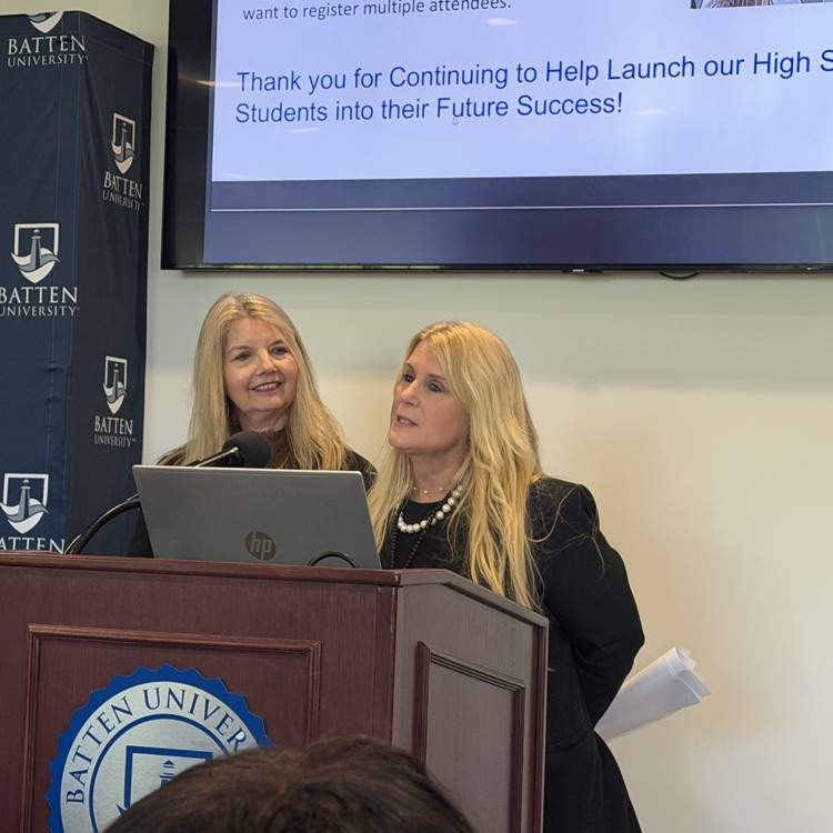 Two women stand at a podium with a microphone and laptop, addressing an audience. Behind them is a large screen with a message.