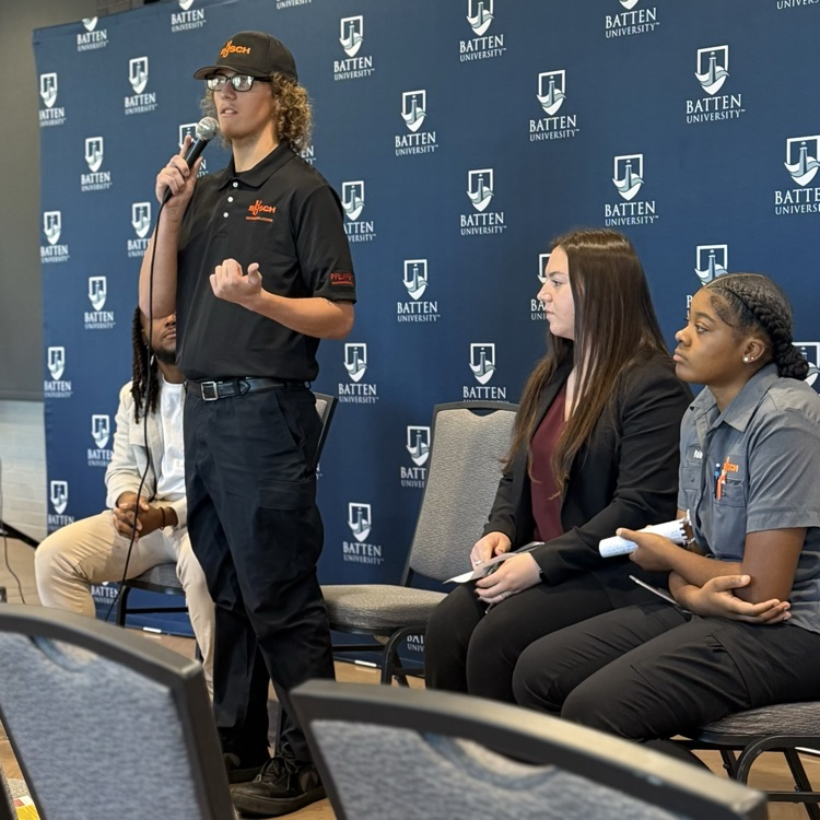 A man with a microphone stands before four seated students. Blue banners with logos are in the background.
