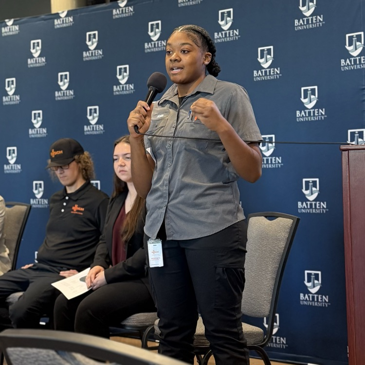 A girl holding a microphone stands before a panel of seated students, against a blue backdrop with logos.