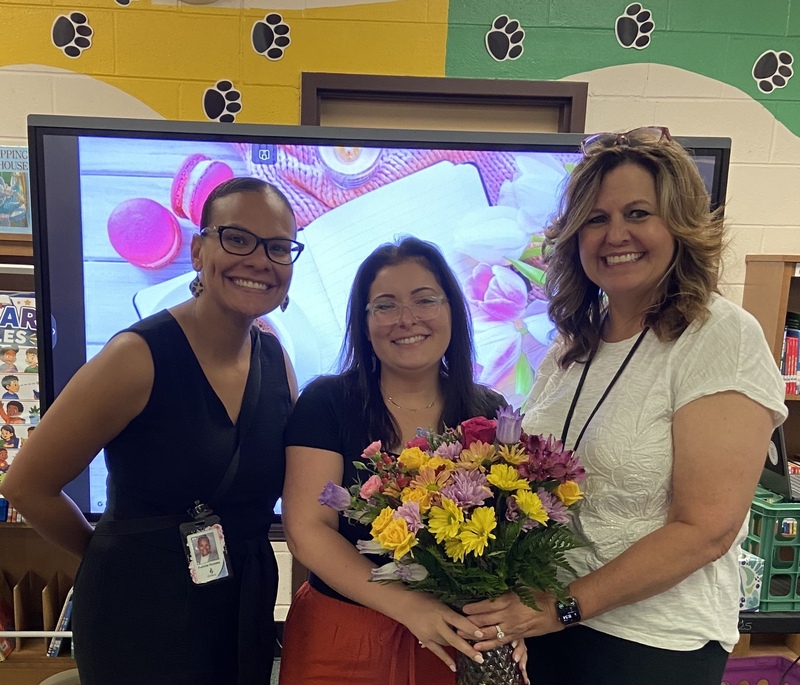 Mrs. Hodge, Greenbrier Primary School’s 2026–2027 Teacher of the Year, stands smiling between two colleagues while holding a colorful bouquet of flowers in the school library. A green and yellow panther paw-print mural decorates the wall behind them.