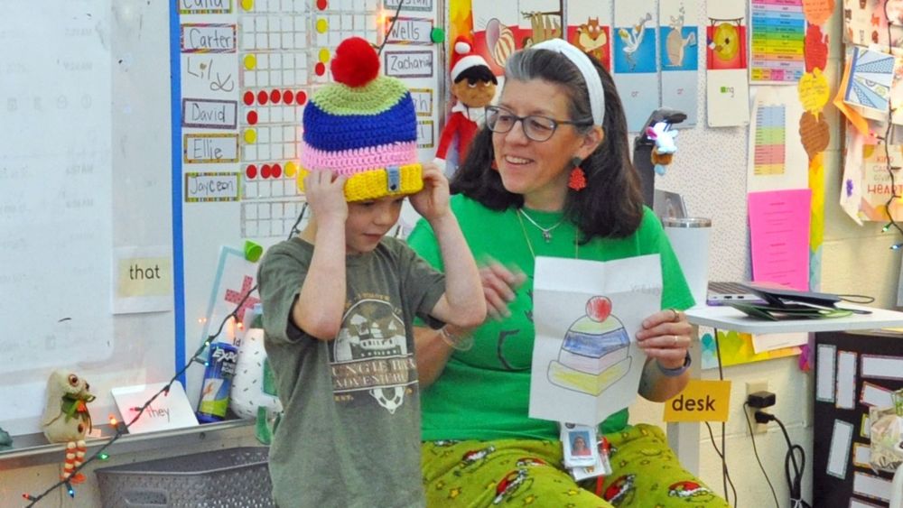 A student wears a colorful crocheted hat that matches a drawing held by their smiling teacher.