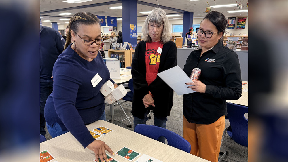 A woman in a navy sweater points to a "Choice A" card on a table while holding small white boxes. Two other women, one in a red t-shirt and another in a black button-down with an orange pair of pants, look on and review a paper document.