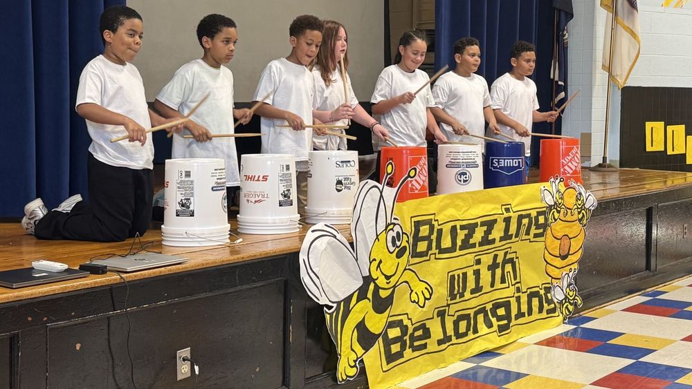 Students playing bucket drums behind a sign that says Buzzing with Belonging