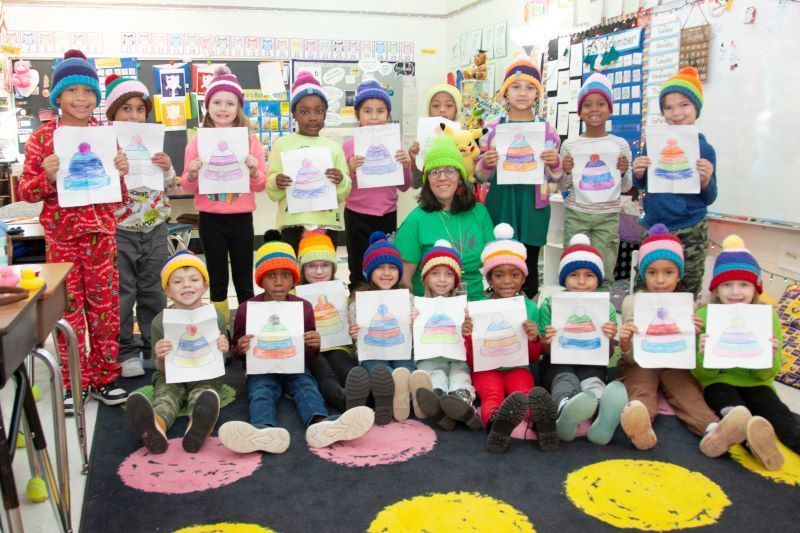 A teacher and a large group of students sit together, each wearing a unique handmade hat that matches a drawing they are holding.
