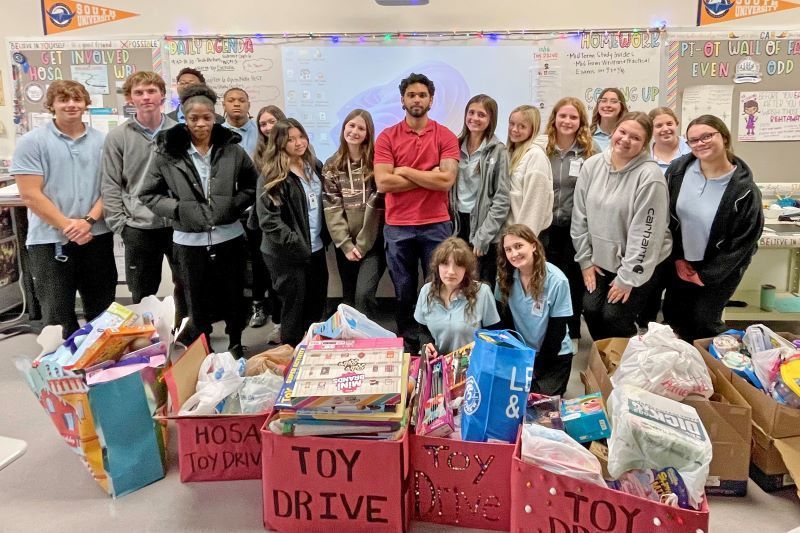 A group of high school students poses behind several boxes overflowing with donated toys for a HOSA toy drive.